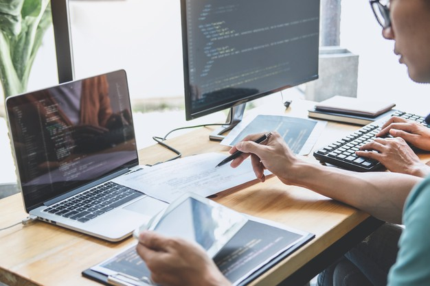 #pracegover: duas pessoas estão em uma mesa com seus computadores executando linhas de programação, um dos usuário está com um notebook e outro está digitando em um computador de mesa.