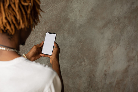 #PraCegoVer: na imagem, um homem está sendo enquadrado de costas e por cima do seu ombro direito. Em suas mãos, ele segura um smartphone.