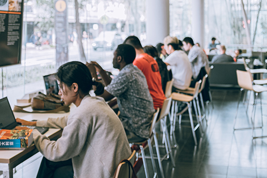 #PraCegoVer: Na imagem, há um cybercafé com vários usuários sentados e digitando em seus respectivos computadores. Ao fundo, há uma vitrine que dá para uma rua arborizada.