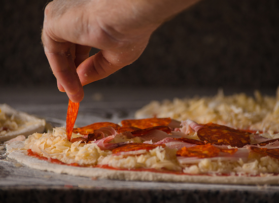 #PraCegoVer:Uma mesa de madeira com três massas de pizzas ainda crus, mas com o recheio já colocado. Na pizza em destaque, uma mão está colocando uma fatia de calabresa nela.