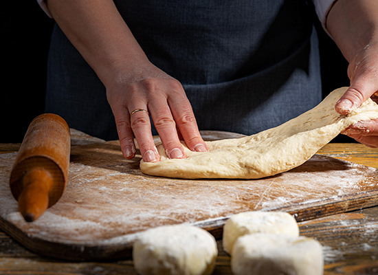 #PraCegoVer: Mesa de madeira com uma tábua e um rolo também de madeira. Em cima da mesa também tem pedaços de massa de pizza e em cima da tábua uma massa sendo aberta por um pizzaiolo.